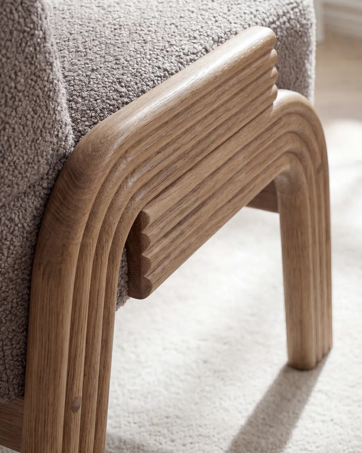 Close-up of a modern upholstered chair corner showing a curved light oak wood frame and textured taupe fabric seat on a light carpet.