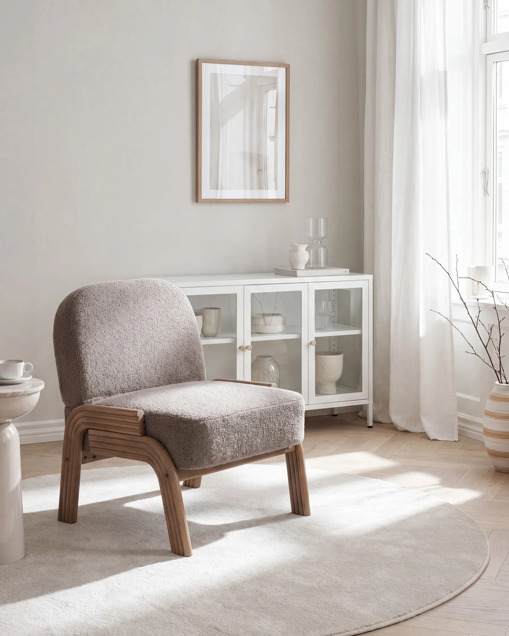 A modern upholstered accent chair with a textured taupe-gray fabric seat and curved light wood frame, positioned in a bright, minimalist room with a white glass cabinet, sheer curtains, and a light rug.
