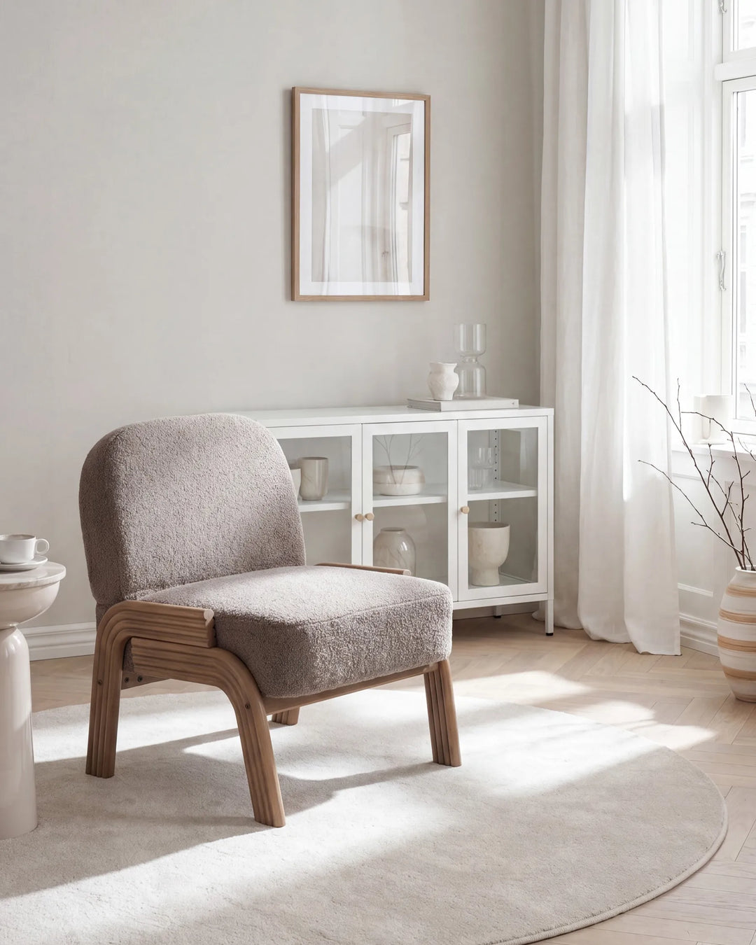 A modern upholstered accent chair with a textured taupe-gray fabric seat and curved light wood frame, positioned in a bright, minimalist room with a white glass cabinet, sheer curtains, and a light rug.