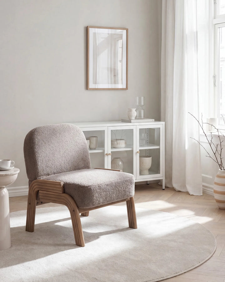 A modern upholstered accent chair with a textured taupe-gray fabric seat and curved light wood frame, positioned in a bright, minimalist room with a white glass cabinet, sheer curtains, and a light rug.
