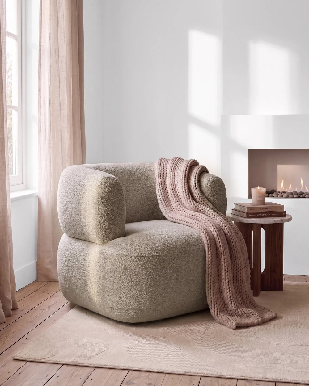 A rounded boucle armchair in a warm light beige tone, styled in a cozy room with a pink knit throw blanket draped over it, a small wooden side table with books and a candle, and a fireplace in the background.