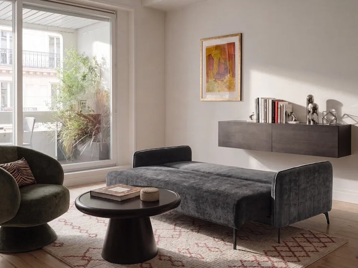 A modern gray velvet chaise lounge bed in a stylishly decorated living room with a dark green armchair, a round black coffee table, and a floating dark gray wall shelf.