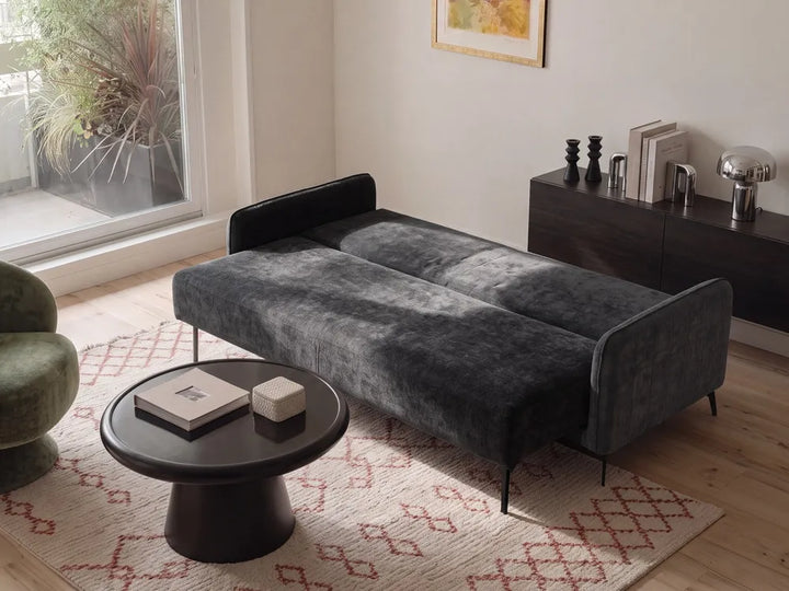 A dark gray velvet daybed with slim black metal legs, styled in a modern living room setting with a round black coffee table, a patterned rug, and a dark wood cabinet in the background.