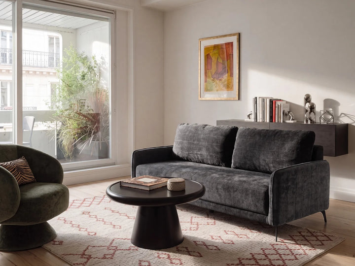 A modern living room featuring a dark gray velvet loveseat sofa, a round black coffee table, a patterned rug, and a large window looking out onto a balcony.