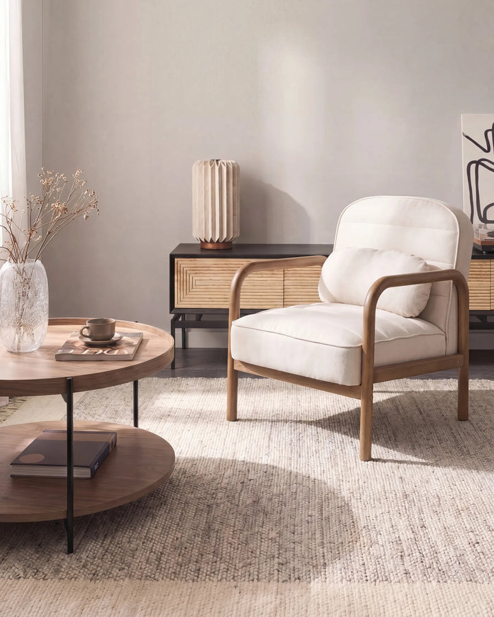 A modern armchair with cream-colored cushions and a light wood frame, styled in a minimalist living room next to a round wooden coffee table and a textured rug.