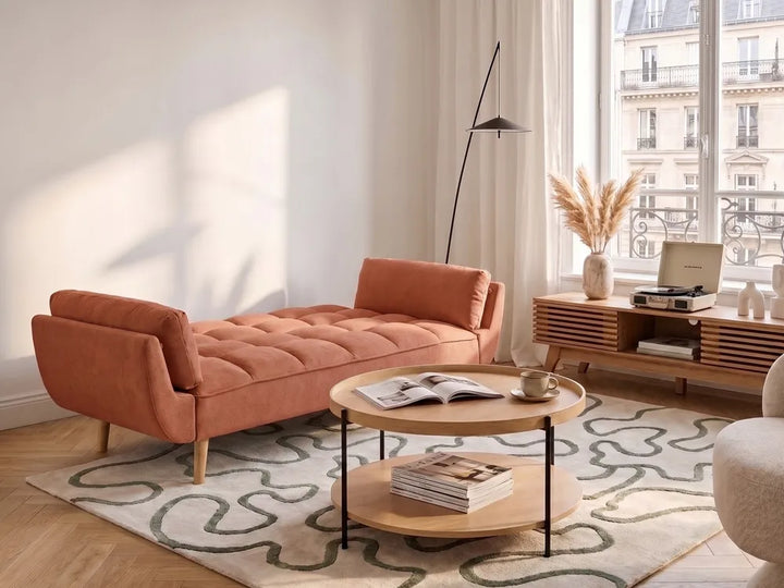 An orange-brown tufted velvet daybed in a modern, sunlit living room with a patterned rug, wooden coffee table, and a large window view.
