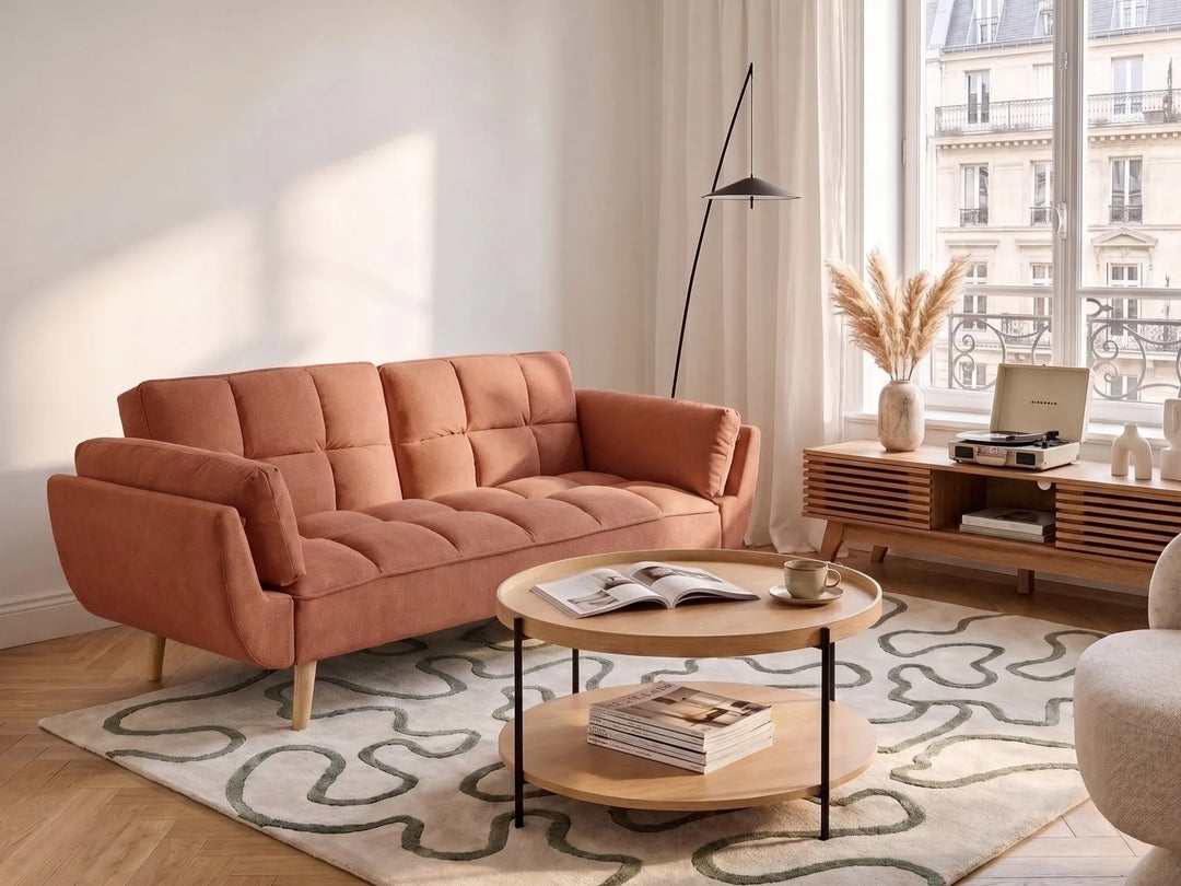 A modern terracotta orange velvet loveseat sofa in a bright, contemporary living room setting with a patterned rug, wooden coffee tables, and a large window.