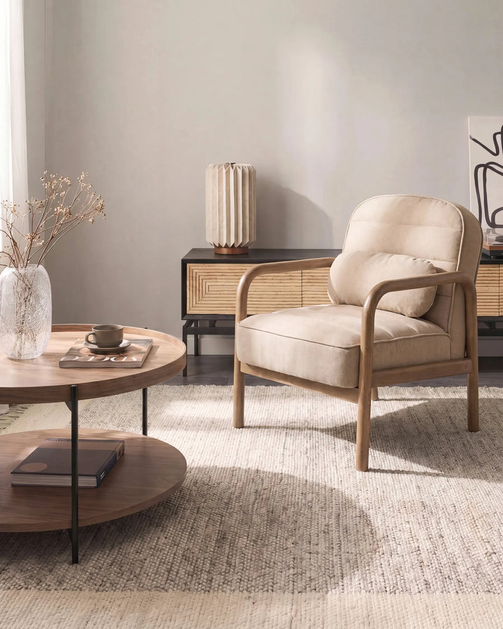A beige upholstered armchair with a light wood frame sits in a styled living room setting next to a round wooden coffee table and a textured rug.