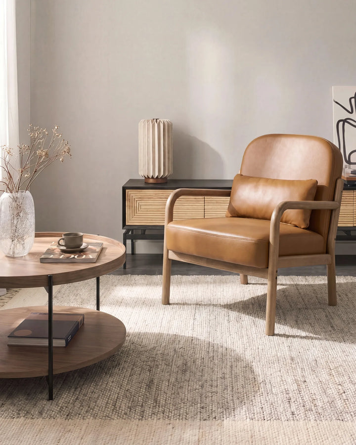 A mid-century modern armchair with tan leather upholstery and a matching lumbar pillow, set in a styled living room next to a round wooden coffee table.