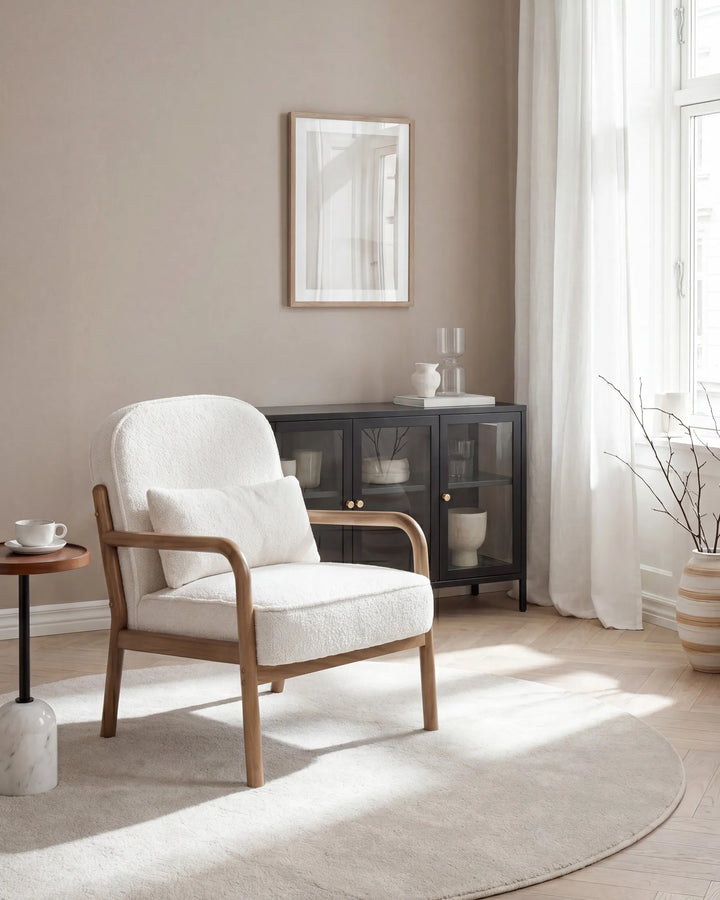 A modern upholstered armchair with a white fabric seat and back, light wood frame, and a matching white throw pillow, placed in a bright, neutral-toned living room next to a small side table.