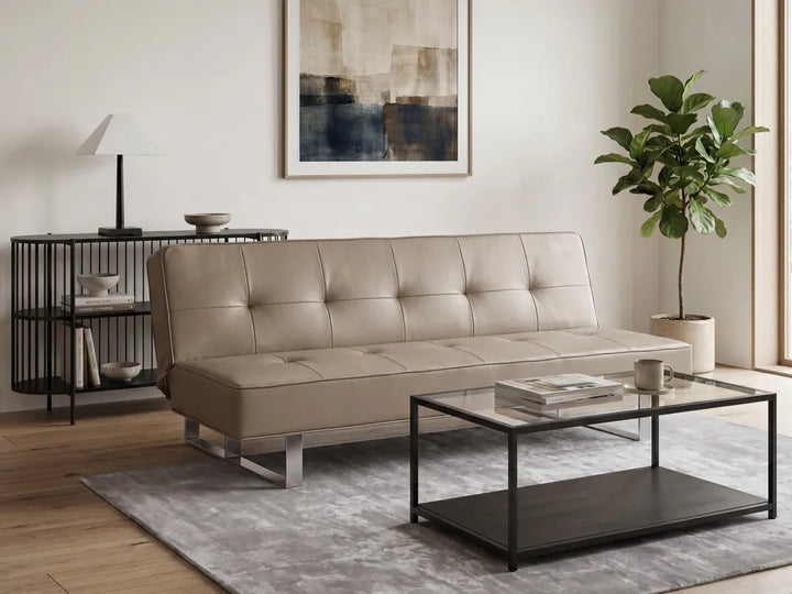 A beige tufted leather sofa in a modern living room setting, styled with a glass-top coffee table, a gray rug, a black side table with a lamp, and a potted plant.