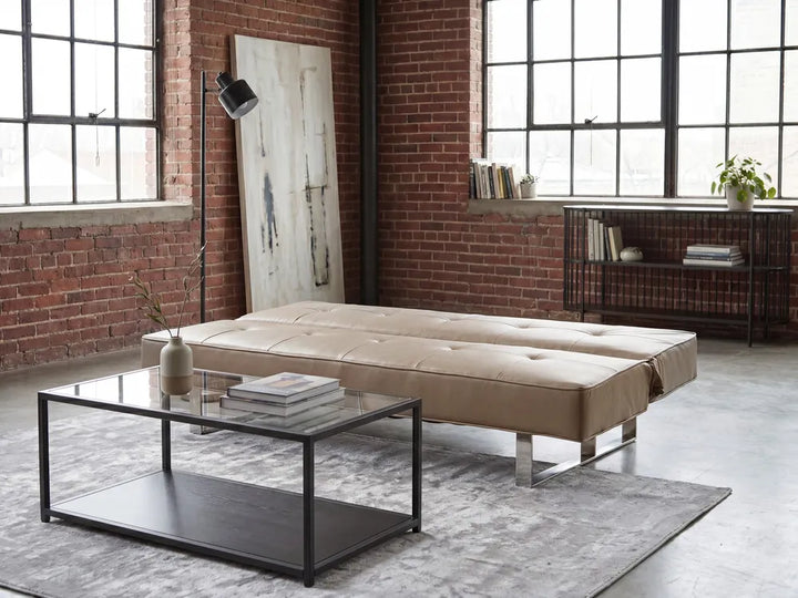 A beige tufted upholstered daybed or chaise lounge with silver metal legs, shown in a modern loft-style room with exposed brick walls, large windows, a glass coffee table, and a gray rug.