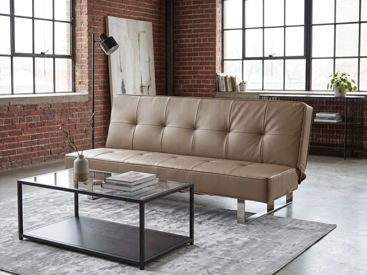 A beige leather futon sofa with tufted button details and silver metal legs, styled in a modern loft room with a glass-top coffee table, gray rug, brick walls, and large industrial windows.