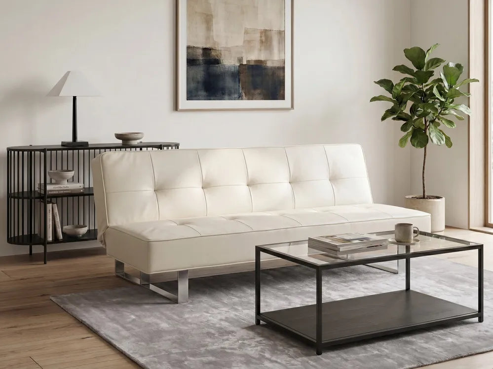 An off-white tufted leather futon sofa with chrome legs, styled in a modern loft room with a glass-top coffee table, gray rug, brick walls, and large industrial windows.
