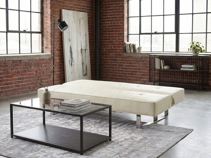 An off-white tufted leather futon sofa with chrome legs, styled in a modern loft room with a glass-top coffee table, gray rug, brick walls, and large industrial windows.