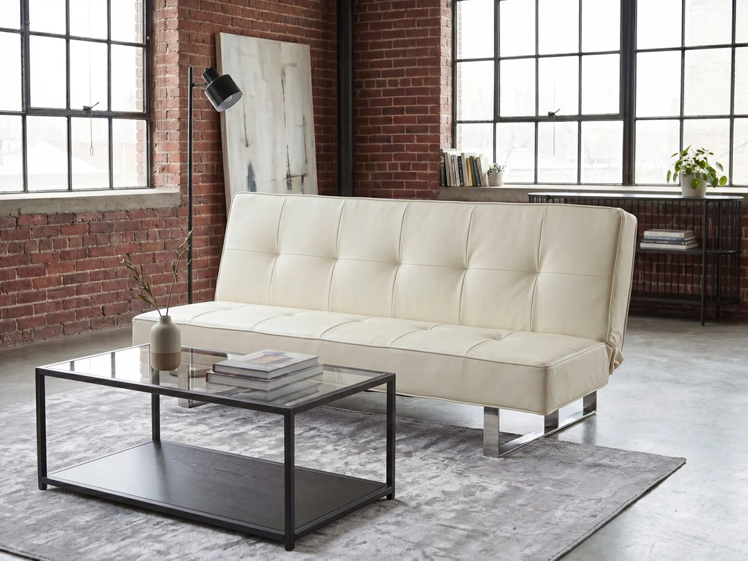 An off-white tufted leather futon sofa with chrome legs, styled in a modern loft room with a glass-top coffee table, gray rug, brick walls, and large industrial windows.