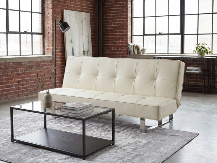 An off-white tufted leather futon sofa with chrome legs, styled in a modern loft room with a glass-top coffee table, gray rug, brick walls, and large industrial windows.