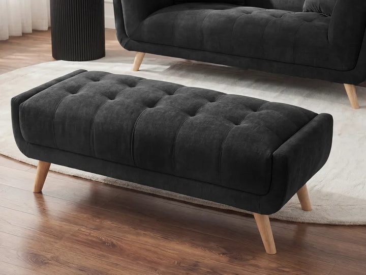 A dark charcoal gray tufted velvet ottoman with light wood legs, shown in a modern living room setting on a white rug with a wooden floor.