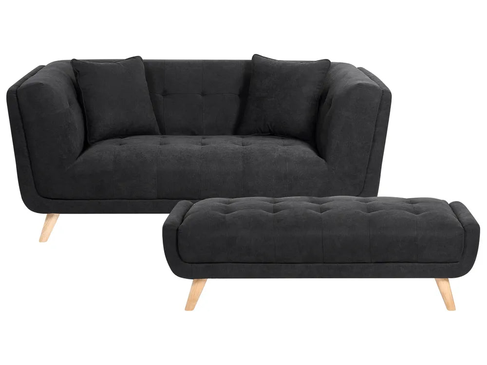 A modern black fabric sofa with matching throw pillows and a matching tufted ottoman, both supported by light natural wood legs, shown on a plain white background.