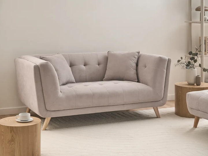 A light beige tufted loveseat sofa with tapered wooden legs, styled in a minimalist living room setting with wooden side tables and neutral decor.