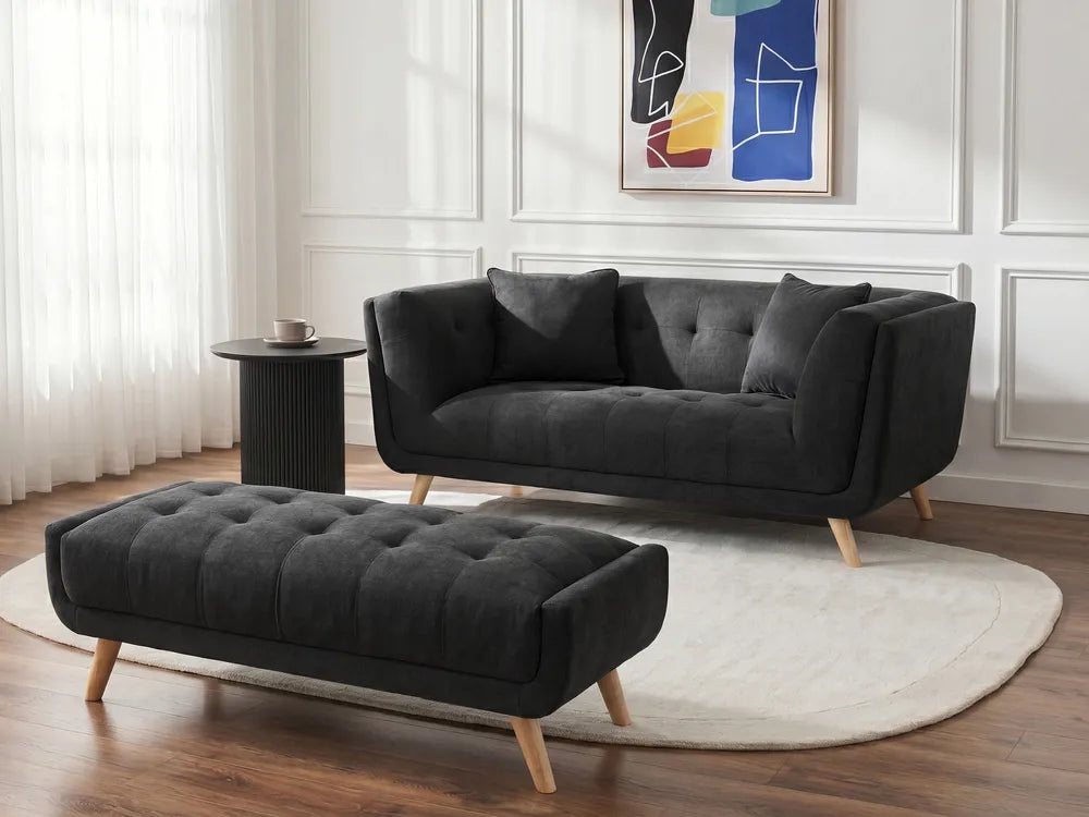A modern black tufted velvet loveseat sofa with light wood legs, paired with a matching black tufted ottoman, set on a round white rug in a bright living room.