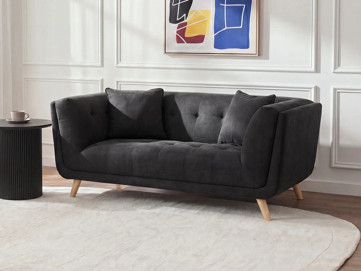 A modern dark gray tufted loveseat with light wood legs, styled in a living room setting with a white paneled wall, abstract art, a white rug, and a black side table.