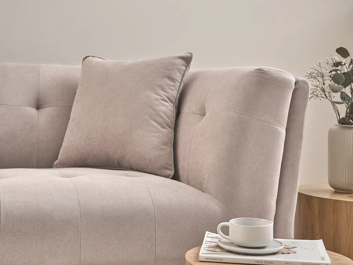 A close-up of a light beige upholstered sofa with a matching throw pillow, shown in a softly styled interior with a wooden side table, a white cup, and a vase with dried stems.