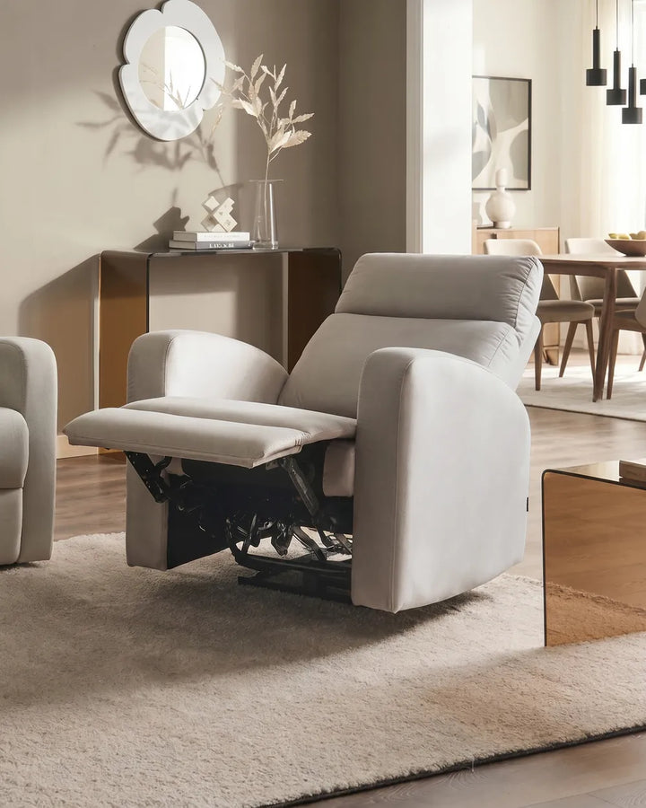 A light beige upholstered recliner chair with the footrest extended, shown in a modern living room setting with a matching rug, sideboard, and warm wood furniture in the background.