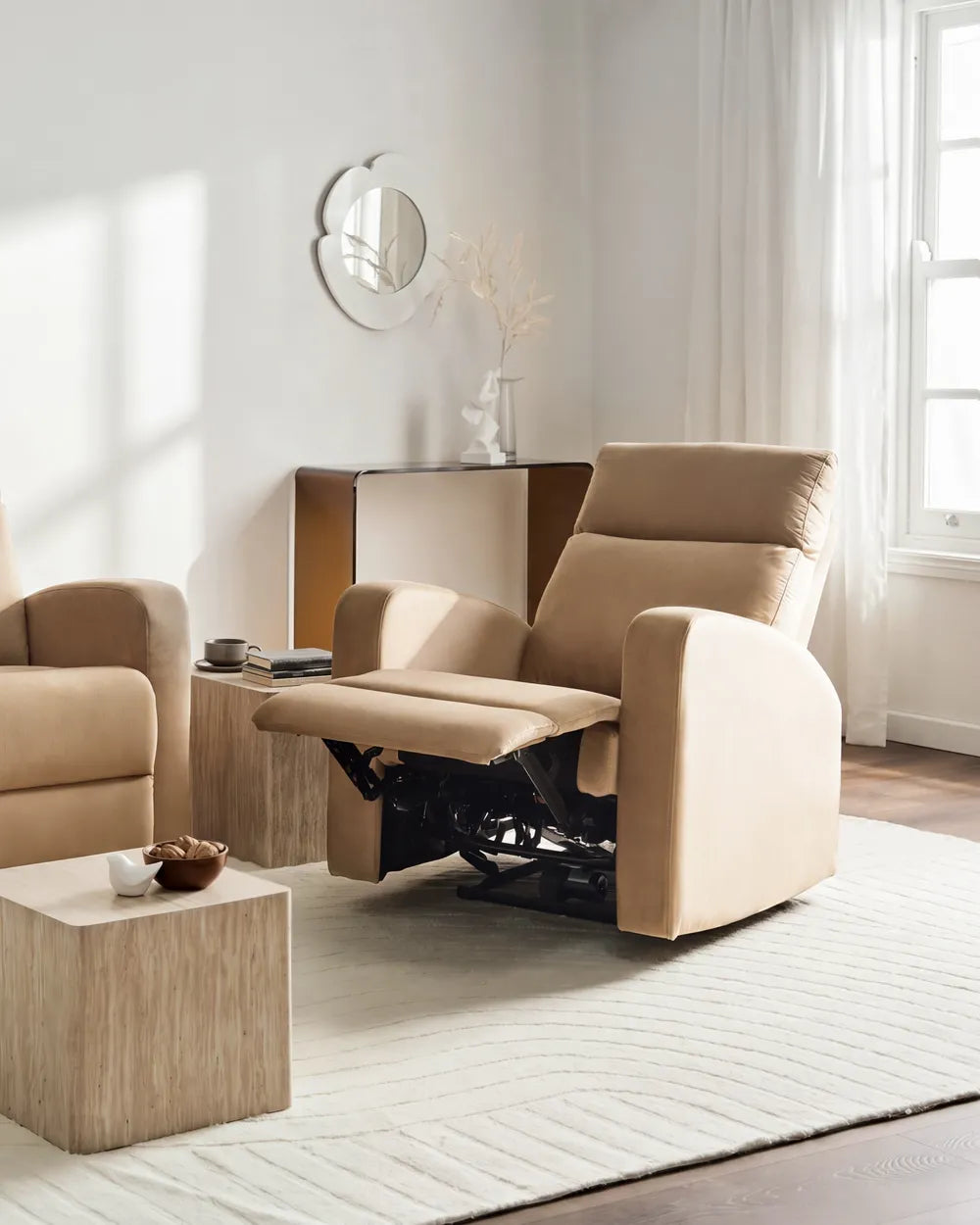A beige upholstered recliner chair with the footrest extended, shown in a bright, modern living room setting with a matching armchair, side table, rug, and window.