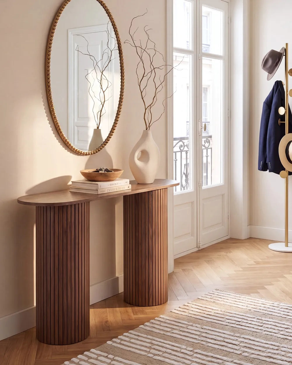 A modern console table with fluted cylindrical legs in a warm brown wood finish, styled in a bright, elegant room with a large oval mirror, white ceramic vases, and a striped rug.