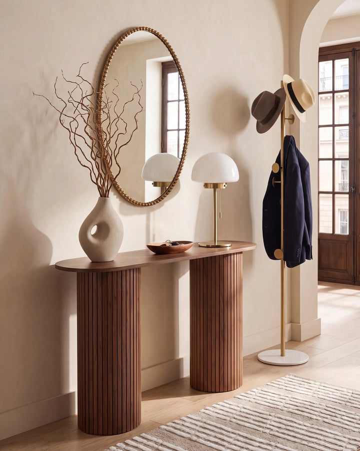 A styled interior entryway featuring a sculptural console table with fluted cylindrical legs, an oval mirror, two white table lamps, a ceramic vase with dried branches, and a gold coat rack holding hats and a navy jacket.