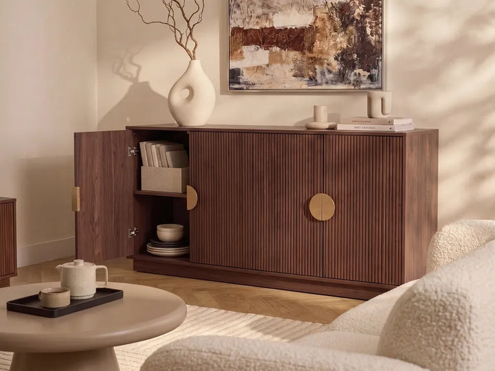 A mid-century modern wooden sideboard cabinet in a warm walnut finish, styled in a cozy living room setting with an open door revealing shelves, brass round handles, and decorative items on top.