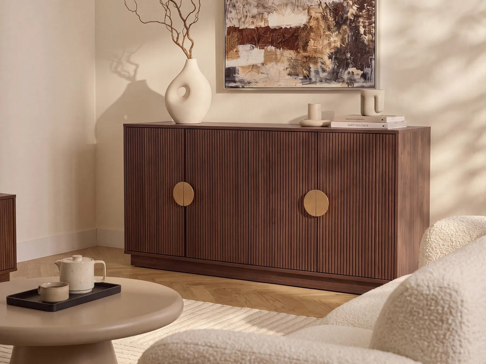 A mid-century modern wooden sideboard cabinet with vertical fluting and round brass handles, styled in a warm, neutral-toned living room next to a round coffee table and textured armchair.