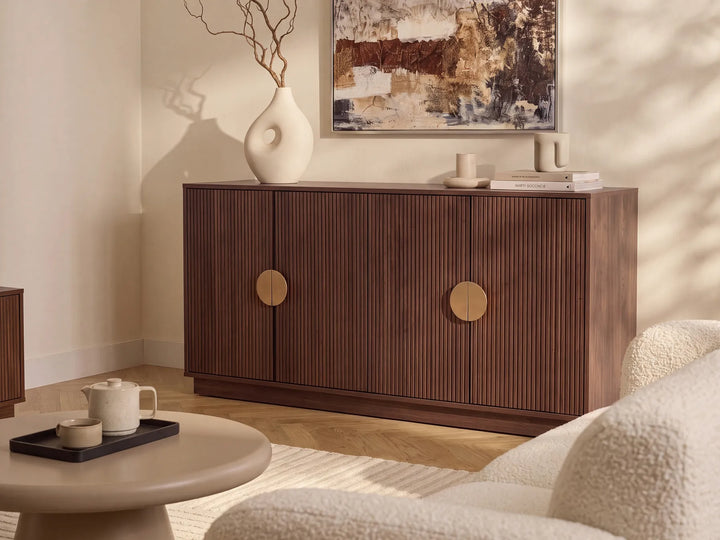 A mid-century modern wooden sideboard cabinet with vertical fluting and round brass handles, styled in a warm, neutral-toned living room next to a round coffee table and textured armchair.
