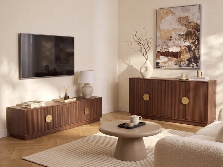 A styled living room featuring two mid-century modern wooden sideboards, one wall-mounted and one freestanding, with brass oval handles. The room includes a beige textured rug, a round coffee table, a wall-mounted TV, and a large abstract painting.