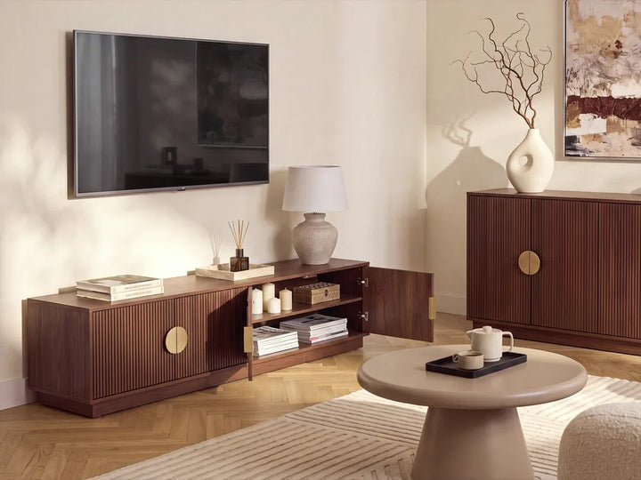 A modern living room scene featuring a dark brown wooden TV stand with vertical fluting and brass circular handles, styled with a wall-mounted TV, a lamp, and books. A matching wooden sideboard and a round beige coffee table are also visible.