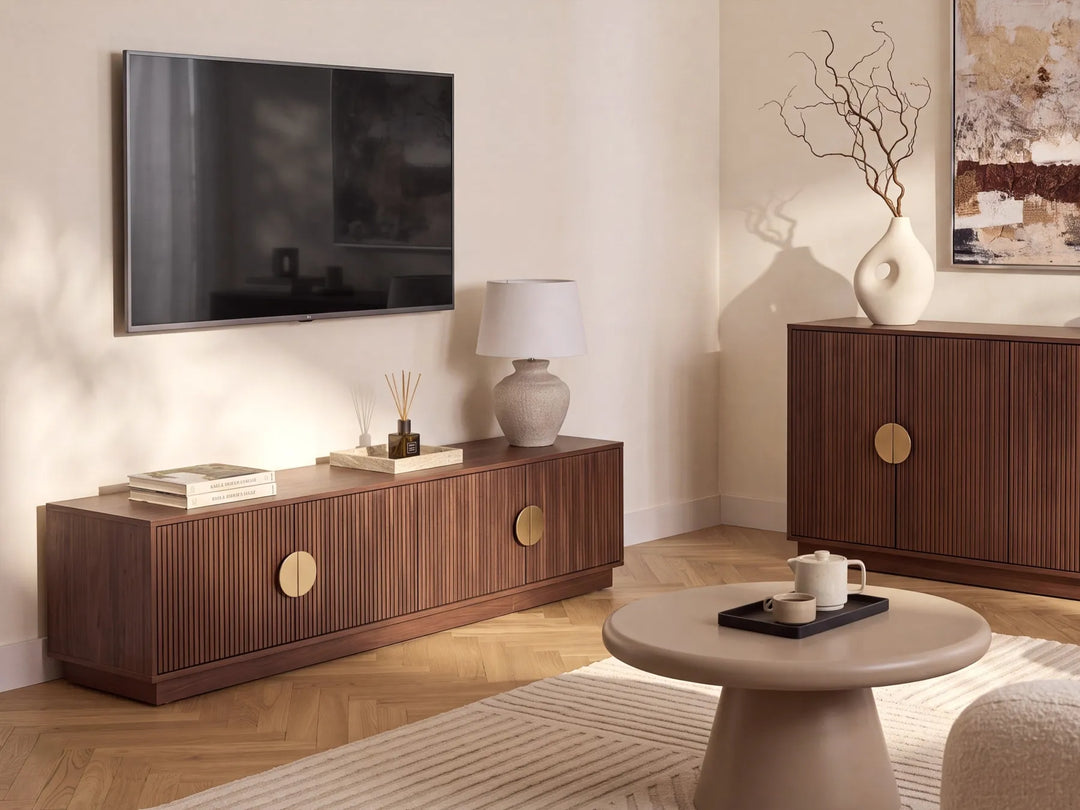 A modern living room featuring a dark brown wood TV stand with fluted vertical panels and round gold-tone pulls, styled with a wall-mounted TV, a ceramic lamp, and decorative items. A matching wooden sideboard and a round beige coffee table are also visible.