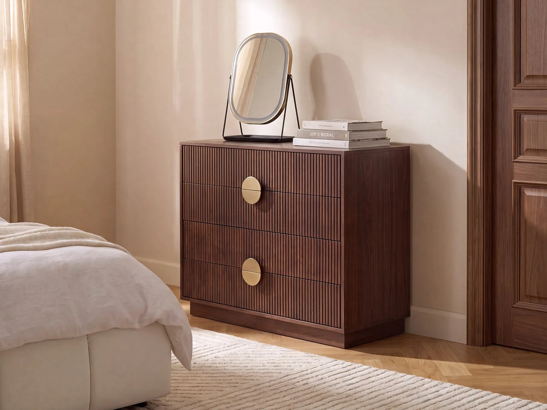 A dark brown wooden nightstand with two drawers featuring vertical slat fronts and round gold-tone handles, styled in a bedroom next to a bed with white bedding. A small oval mirror and books sit on top.