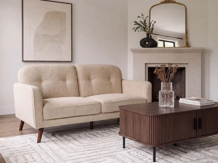 A cream-colored tufted loveseat sofa in a modern living room setting, positioned next to a dark wood coffee table and in front of a fireplace.