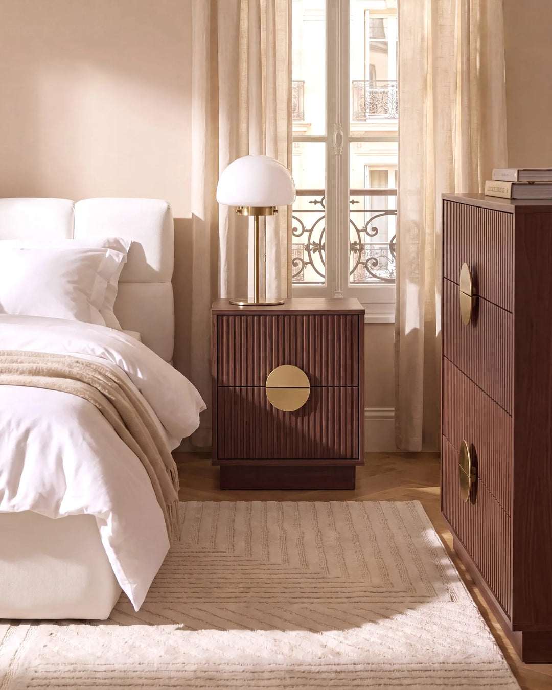 A modern bedroom scene featuring a dark brown ribbed nightstand with a round gold pull, styled with a white bed, a white table lamp, and a matching dark brown chest of drawers with gold handles.