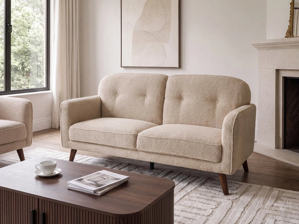 A beige upholstered loveseat with tufted back cushions and tapered dark wood legs, shown in a bright living room with a coffee table, window, and fireplace.