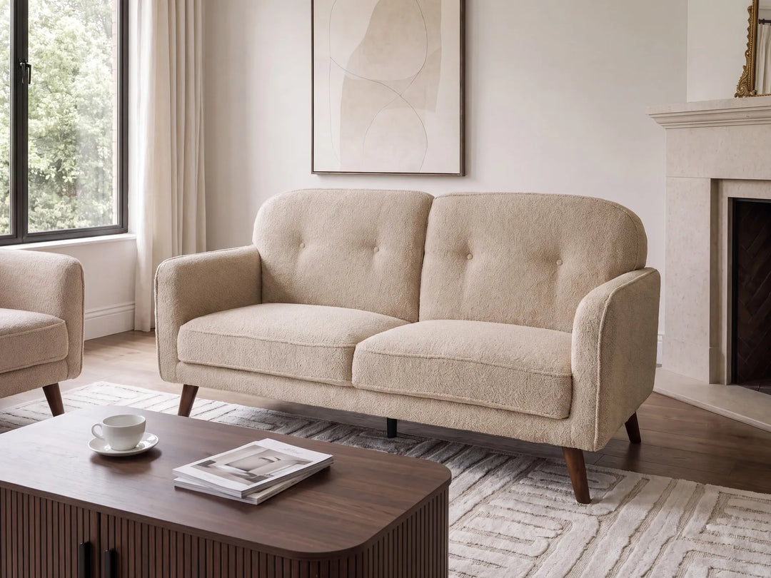 A beige upholstered loveseat with tufted back cushions and tapered dark wood legs, shown in a bright living room with a coffee table, window, and fireplace.