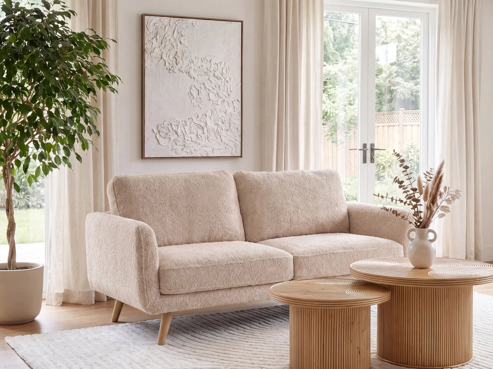 A beige bouclé loveseat sofa in a bright, modern living room setting with a wooden coffee table, a potted plant, and a large window.