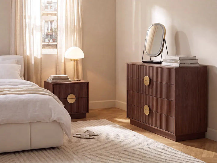 A styled bedroom scene featuring a dark brown ribbed wood chest of drawers with round gold-tone handles, alongside a matching nightstand and a bed with white bedding.