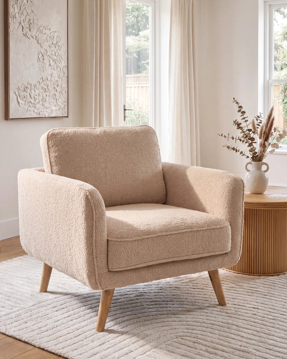 A modern beige bouclé armchair with tapered wooden legs, styled in a bright, neutral-toned living room next to a wooden side table and window.