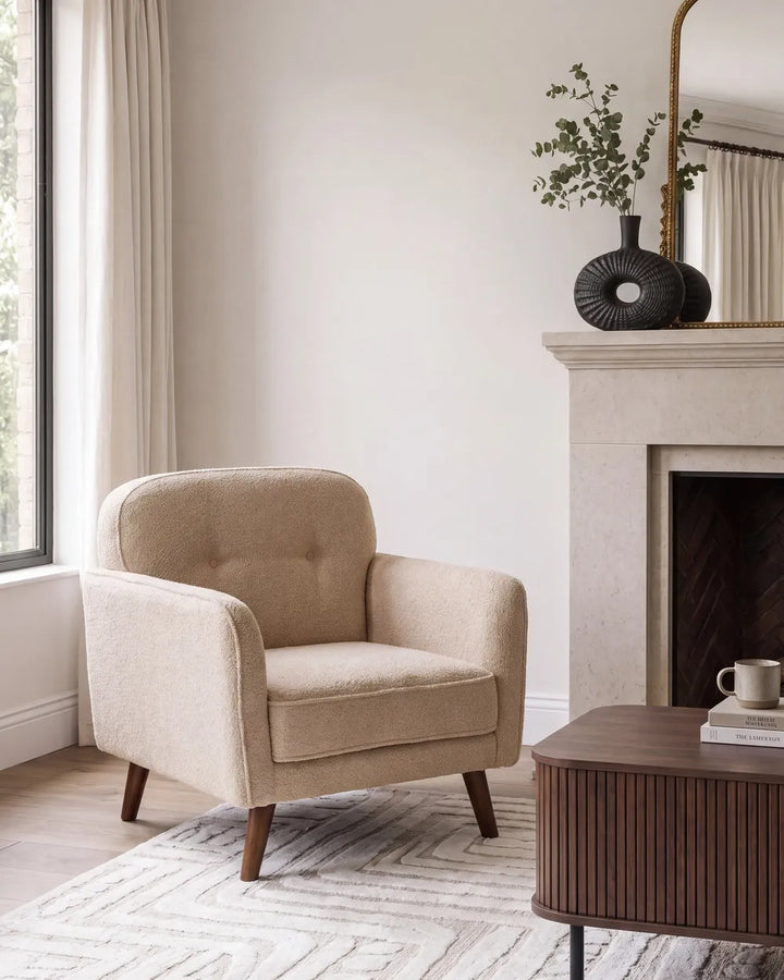 A beige upholstered armchair with tufted backrest and tapered wooden legs, placed in a bright living room beside a fireplace and window.