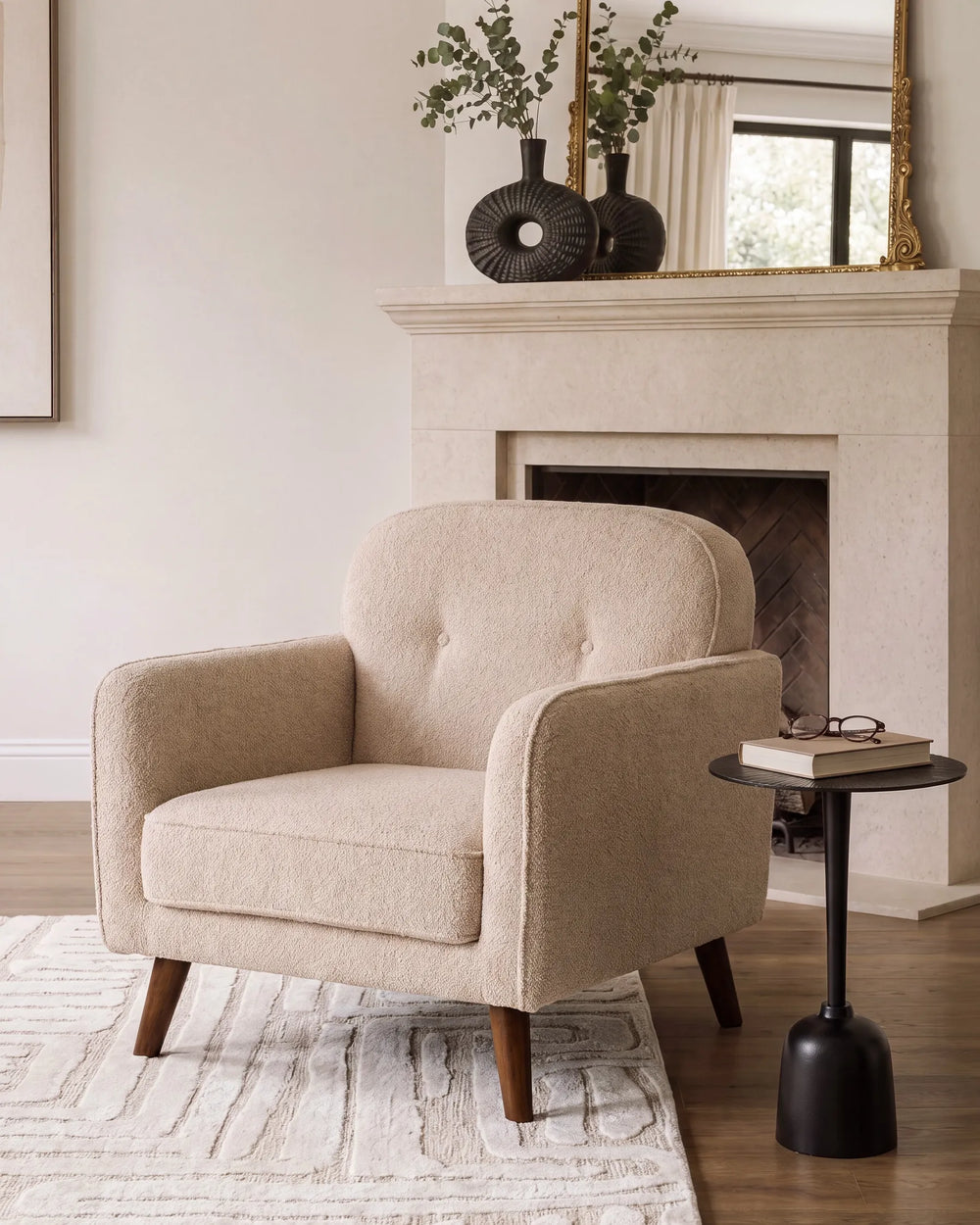 A beige upholstered armchair with tufted backrest and tapered wooden legs, styled in a living room setting next to a black side table in front of a fireplace.