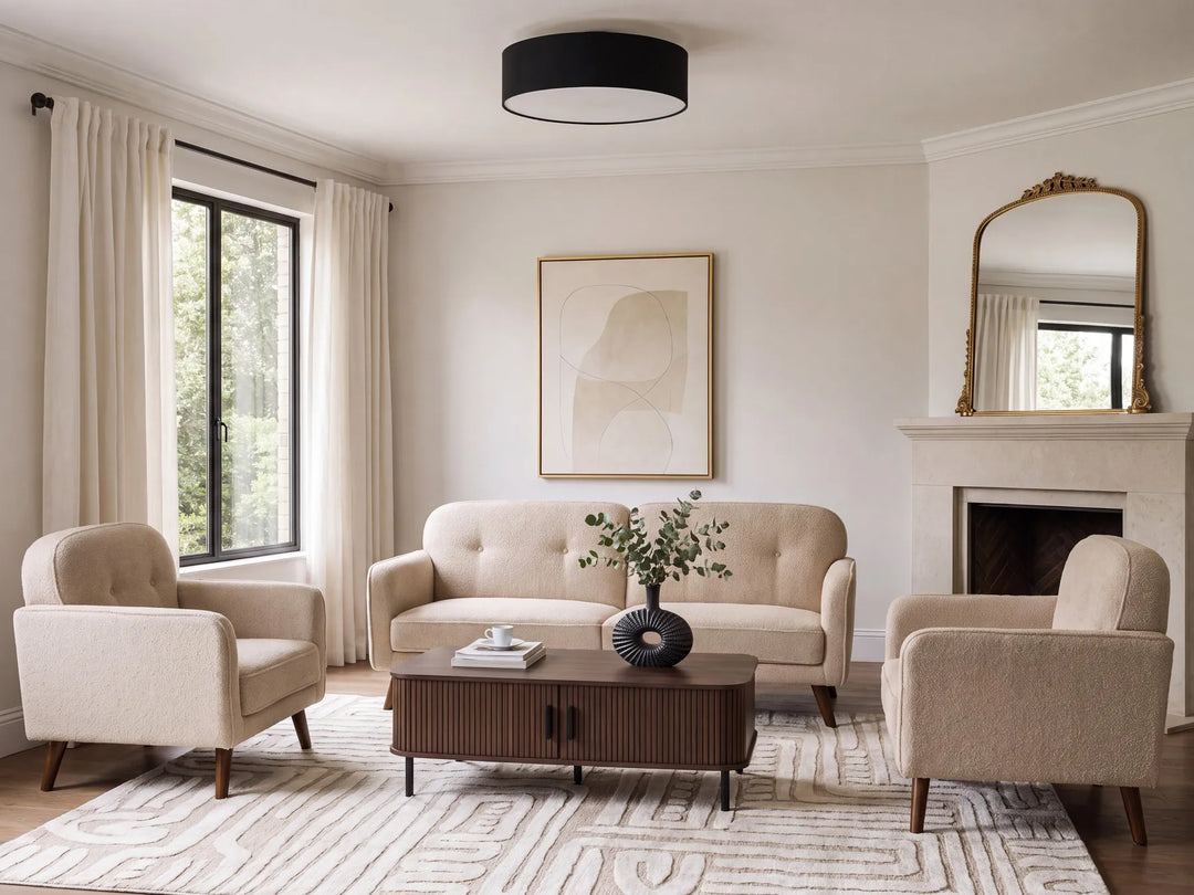 A styled living room featuring a beige tufted sofa, two matching armchairs, a dark wood coffee table, and a patterned rug, set against cream walls with a fireplace, window with curtains, and a black drum ceiling light.