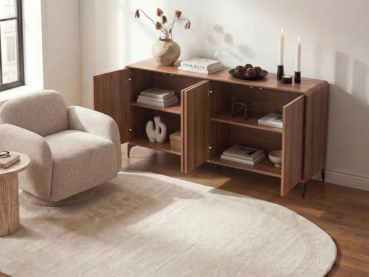 A mid-century modern wooden credenza or sideboard cabinet with open doors, styled in a bright living room next to a boucle armchair and a round cream rug.