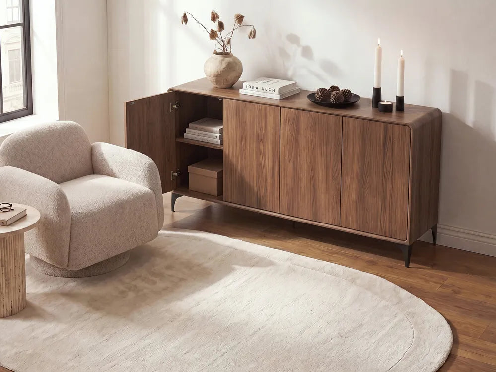 A mid-century modern wooden sideboard cabinet in a warm walnut finish, styled in a bright living room next to a beige armchair and a cream rug.